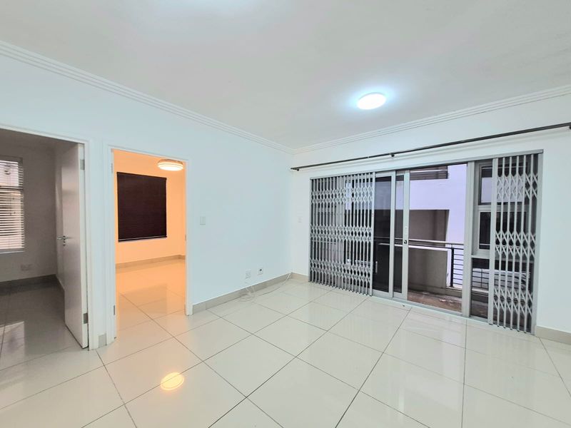 A bright, empty living area featuring neutral tiled flooring, sliding glass doors with security gates, and access to an adjoining room.