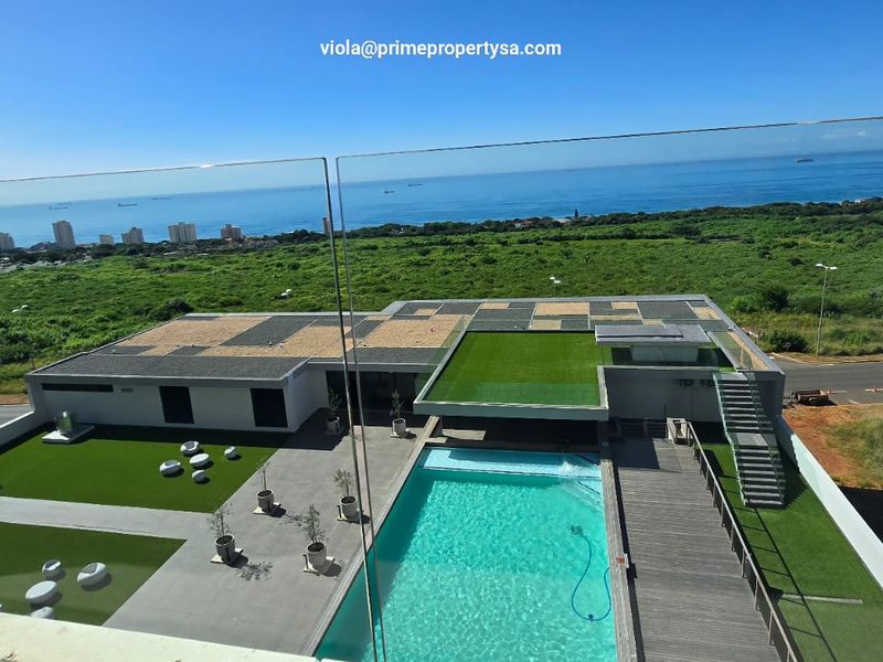 Elevated view overlooking a modern apartment complex featuring a swimming pool, landscaped artificial lawn areas, and a scenic coastal horizon.