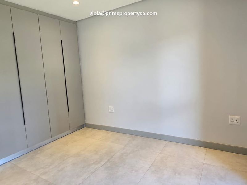 A bright, modern bedroom featuring neutral tiled flooring, built-in cupboards with sleek handles, and large aluminium-framed windows.