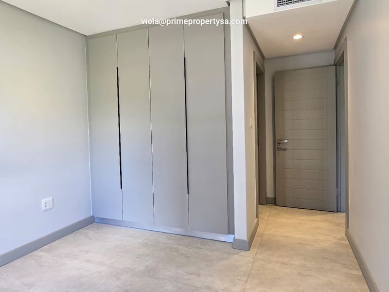 A modern, empty bedroom featuring neutral tiled flooring, built-in cupboards with sleek handles, and recessed ceiling lighting.