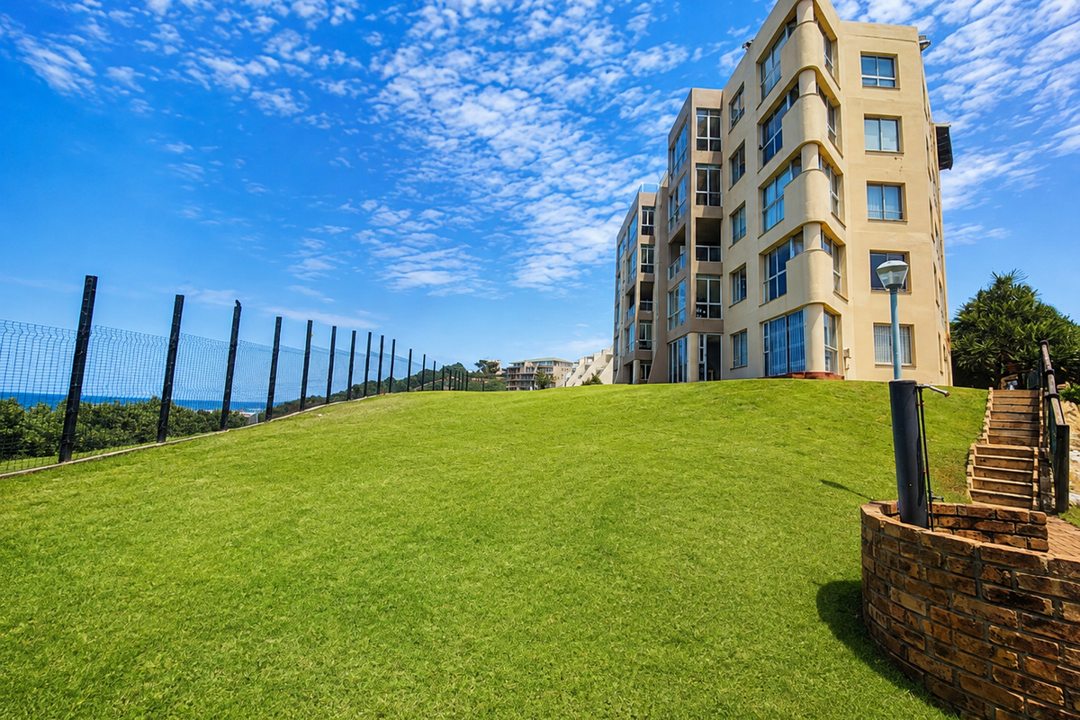 Exterior view of a multi-story apartment building featuring a well-maintained lawn, brick retaining wall, and perimeter security fencing.