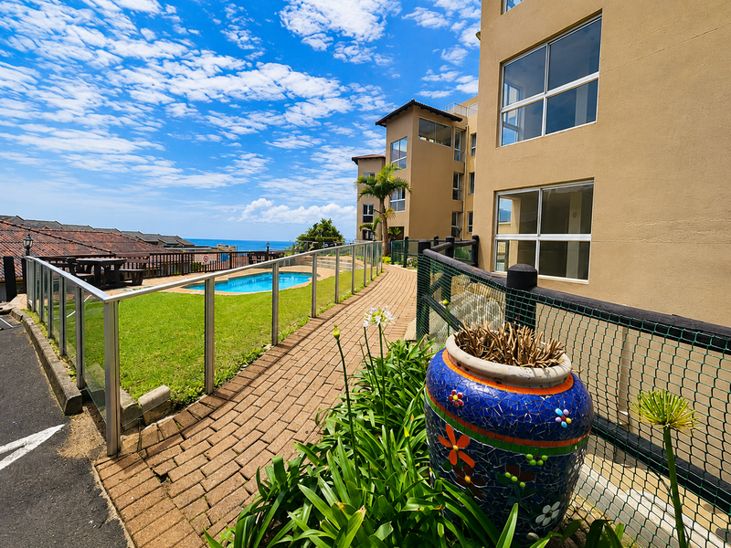 Exterior view of an apartment complex featuring a swimming pool, paved walkway, and landscaped garden area with ocean views.