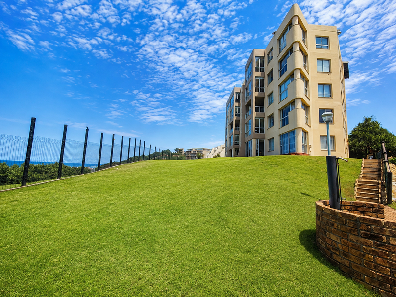 Exterior view of a multi-story apartment building featuring a well-maintained lawn, brick retaining wall, and perimeter security fencing.