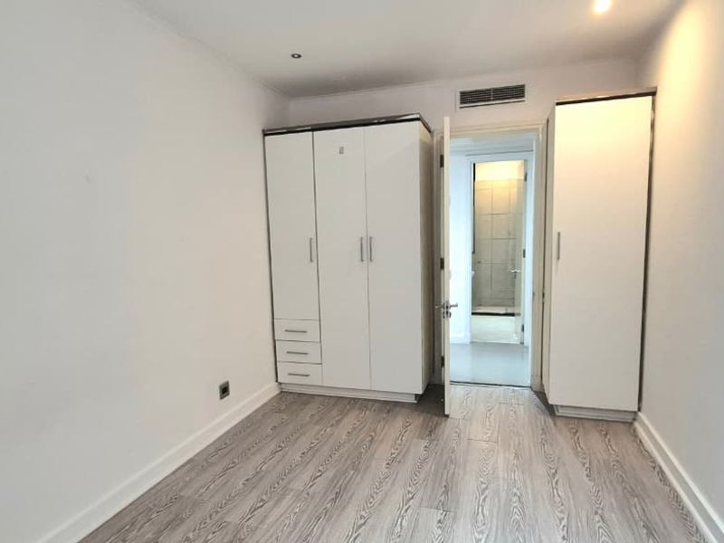 A bright, empty bedroom featuring modern laminate flooring, built-in white wardrobes, and integrated air conditioning, offering a clean and neutral space.