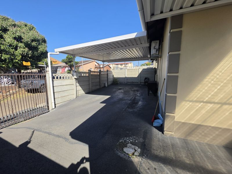 A paved driveway area featuring a covered carport, enclosed by precast concrete boundary walls and a metal entrance gate.
