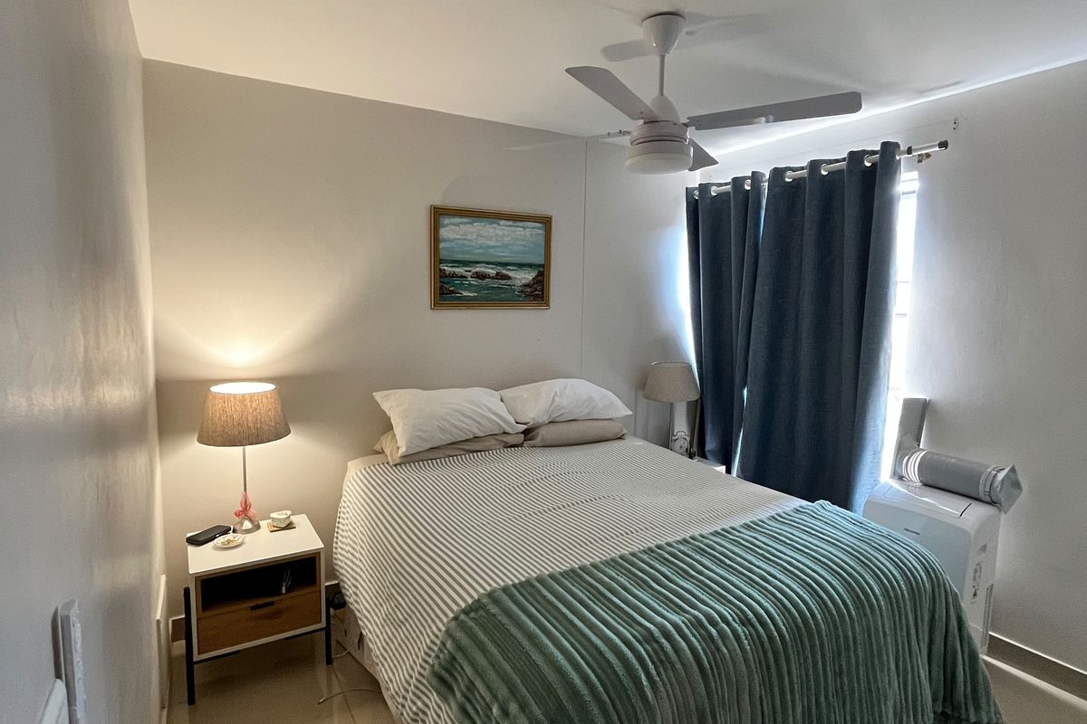 A well-maintained bedroom featuring neutral tiled flooring, a ceiling fan, and large windows with curtains, providing a comfortable and functional living space.