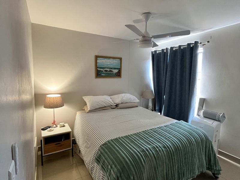 A well-maintained bedroom featuring neutral tiled flooring, a ceiling fan, and large windows with curtains, providing a comfortable and functional living space.