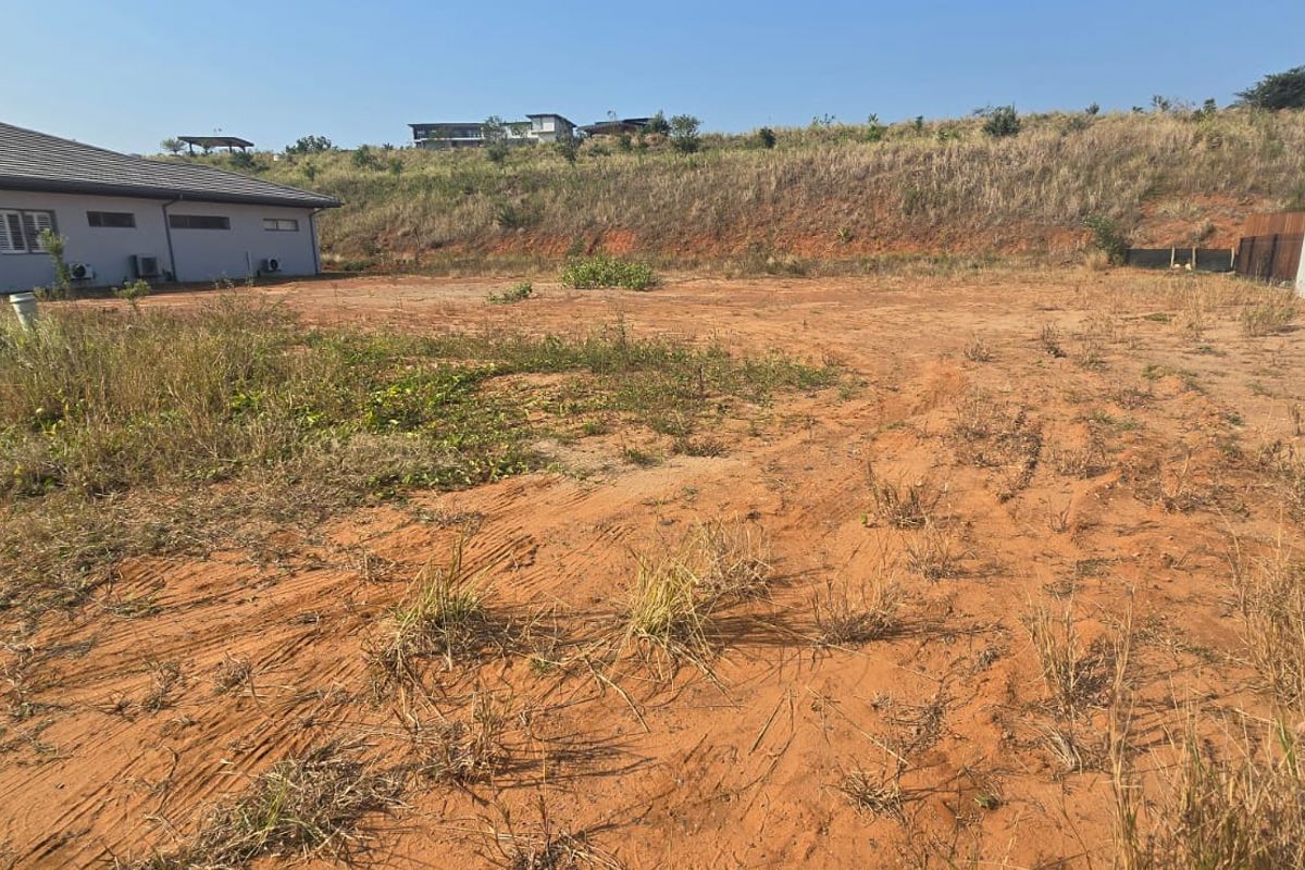 A vacant residential plot featuring natural earth terrain and surrounding boundary fencing, situated adjacent to existing residential structures.