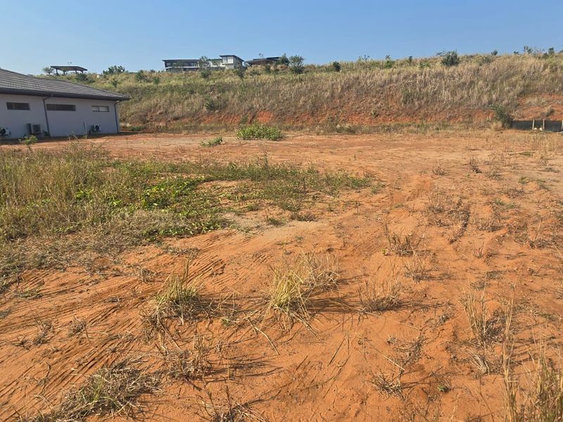 A vacant residential plot featuring natural earth terrain and surrounding boundary fencing, situated adjacent to existing residential structures.