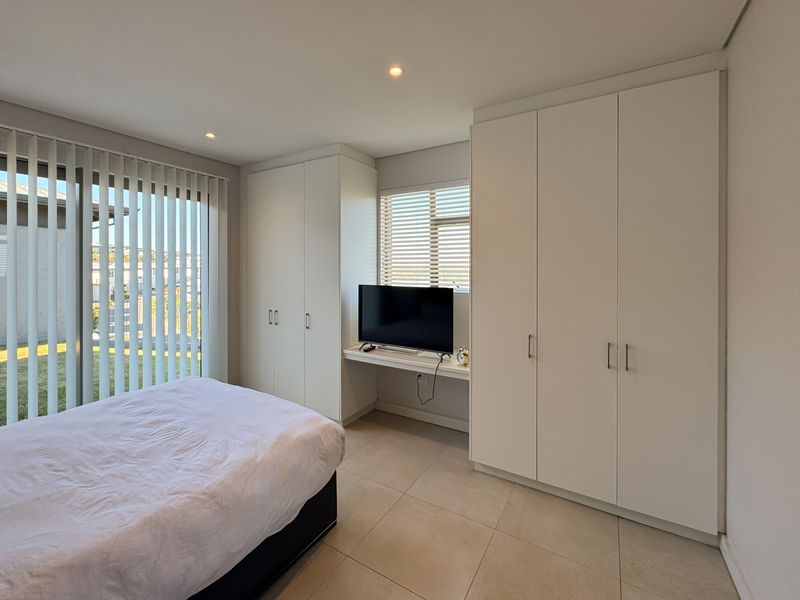 A modern bedroom featuring extensive built-in cabinetry, a dedicated desk area, tiled flooring, and large windows with integrated blinds for natural light control.