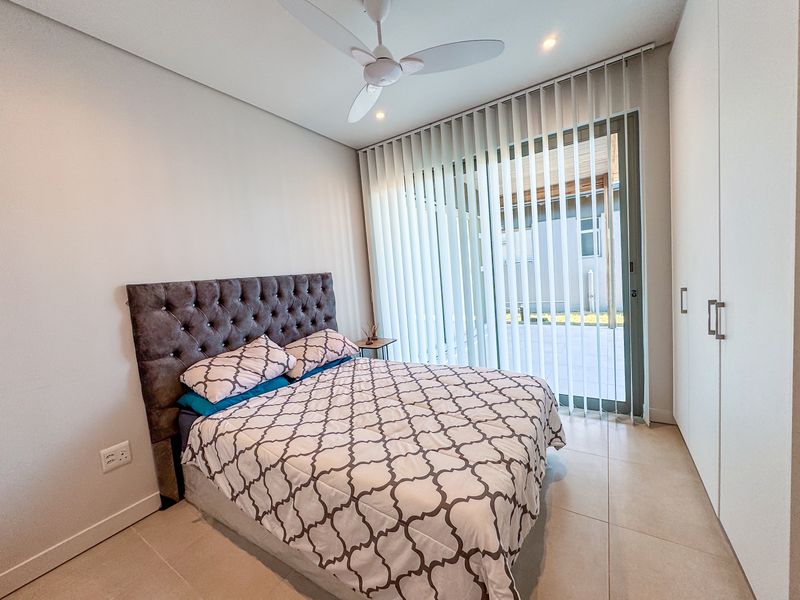 A modern bedroom featuring tiled flooring, built-in cupboards, a ceiling fan, and a sliding glass door with vertical blinds leading to the exterior.