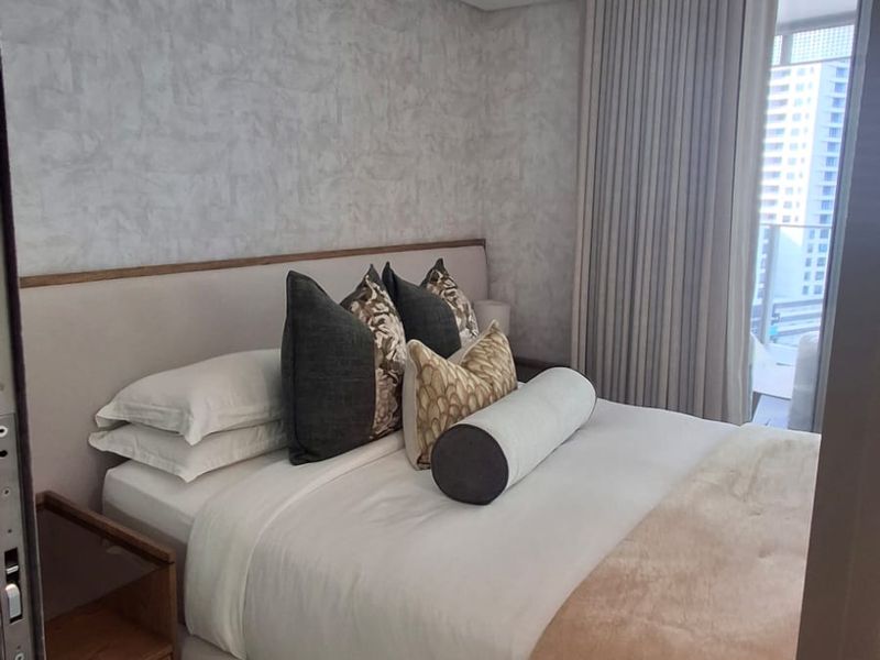 A modern, well-maintained bedroom featuring a built-in headboard, neutral textured wallpaper, and floor-to-ceiling curtains with access to a private balcony.