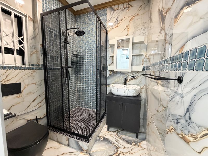 Modern bathroom featuring a walk-in shower with black frame, vessel sink vanity, wall-mounted toilet, and decorative marble-effect wall and floor tiling.