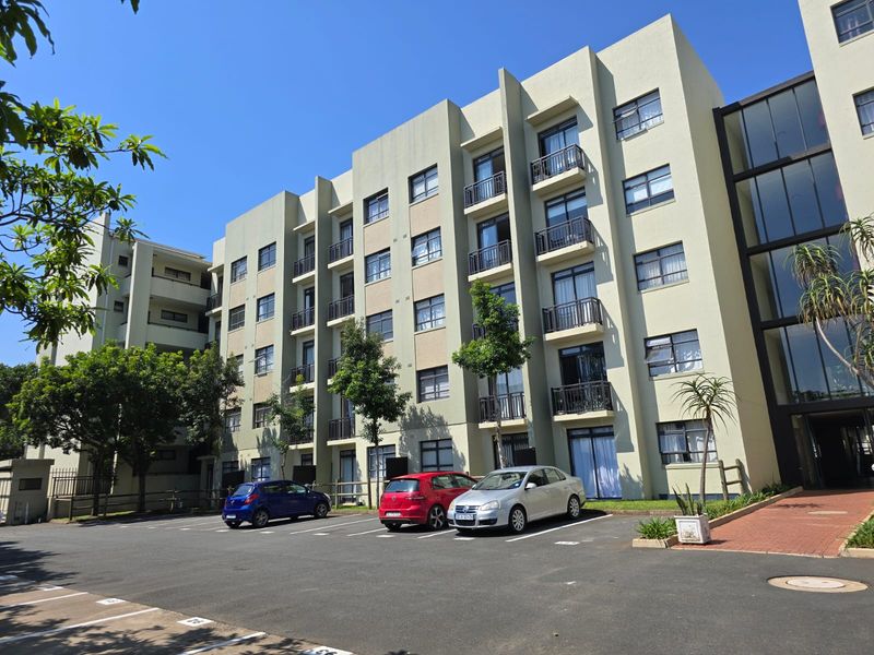 A multi-story apartment building exterior featuring a paved parking area, balconies with railings, and large glass sliding doors.