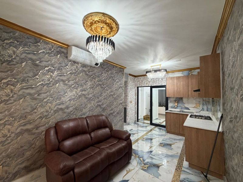 Open-plan living and kitchen area featuring marble-effect finishes, decorative gold crown molding, crystal chandeliers, and built-in kitchen cabinetry with a gas hob.