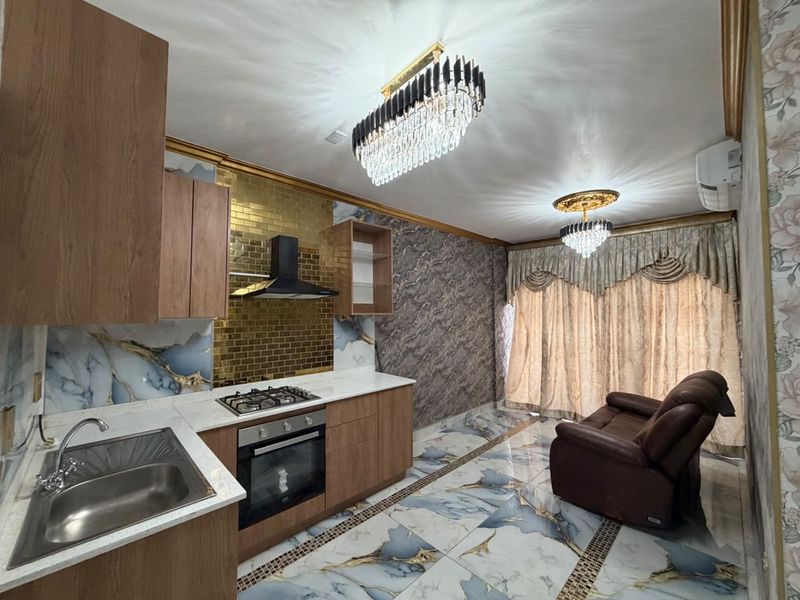 Modern open-plan kitchen featuring wood-finish cabinetry, stone countertops, integrated appliances, and decorative lighting, seamlessly flowing into a living area with marble-effect flooring.