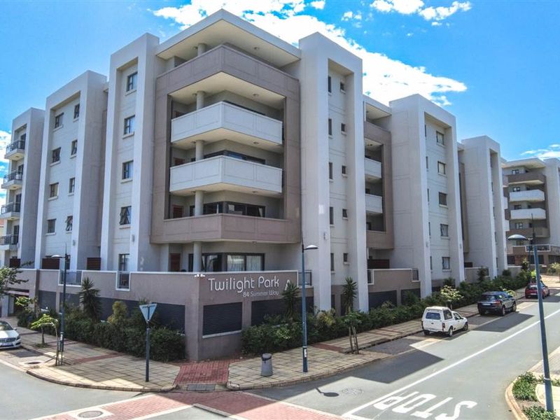 A modern multi-story apartment complex exterior featuring balconies and a secure perimeter wall, situated on a corner lot.