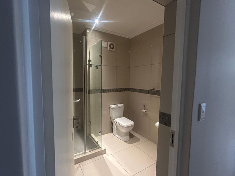 A clean, modern bathroom featuring a glass-enclosed shower, neutral wall and floor tiling, and integrated extractor fan.