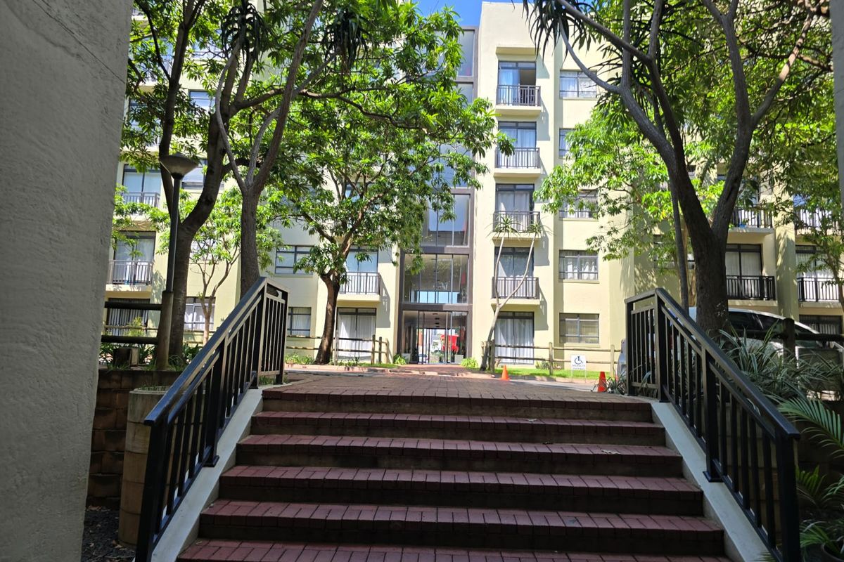 A view of the apartment building exterior featuring a brick staircase leading to the main entrance, surrounded by mature trees and landscaped grounds.