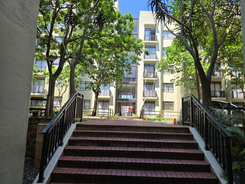 A view of the apartment building exterior featuring a brick staircase leading to the main entrance, surrounded by mature trees and landscaped grounds.