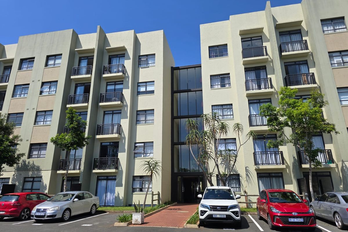 Front elevation of a multi-story apartment complex featuring balconies, a central glass stairwell, and an adjacent paved parking area.