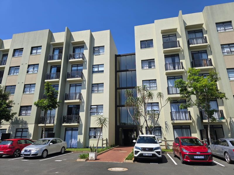 Front elevation of a multi-story apartment complex featuring balconies, a central glass stairwell, and an adjacent paved parking area.