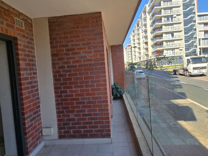 A modern balcony featuring a glass balustrade, exposed brick wall detailing, and tiled flooring, offering an open view of the surrounding apartment complex.