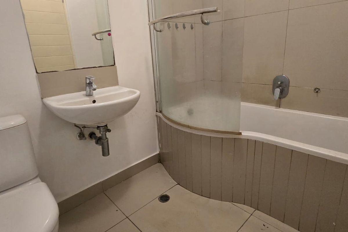 A compact bathroom featuring a pedestal basin, toilet, and a curved bathtub with a glass shower screen, requiring cosmetic updates to finishes.