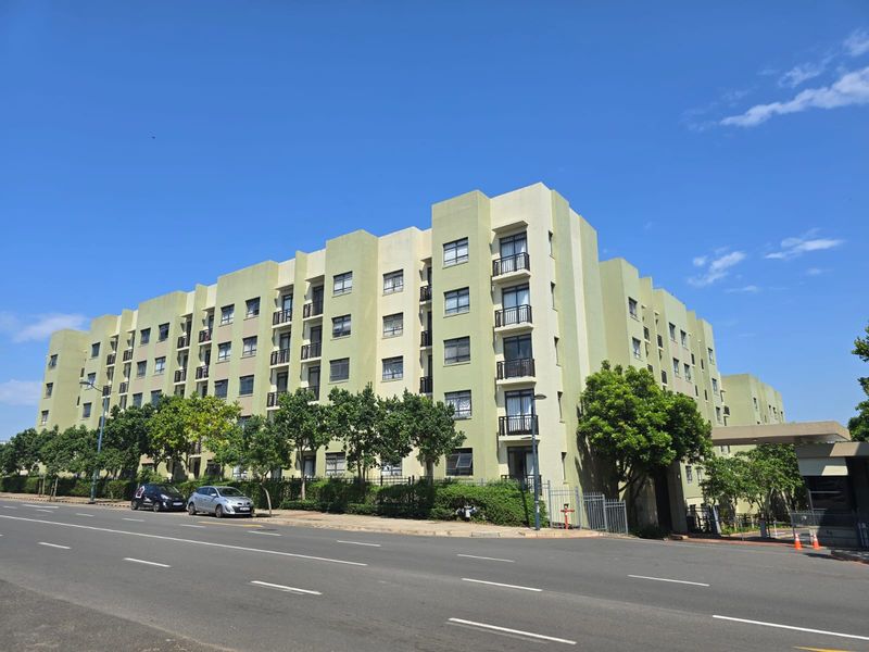 A clear street-level view of the apartment complex facade, featuring multiple balconies and established landscaping along the front boundary.
