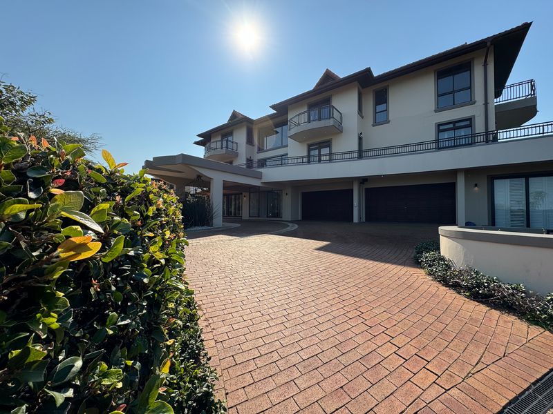 A grand multi-story residence featuring a spacious brick-paved driveway, covered entrance, and multiple balconies under a clear sky.