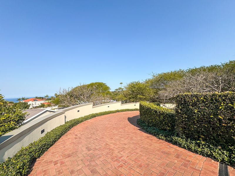 A well-maintained brick paved pathway winds through landscaped gardens, featuring integrated retaining walls and exterior lighting with distant coastal views.