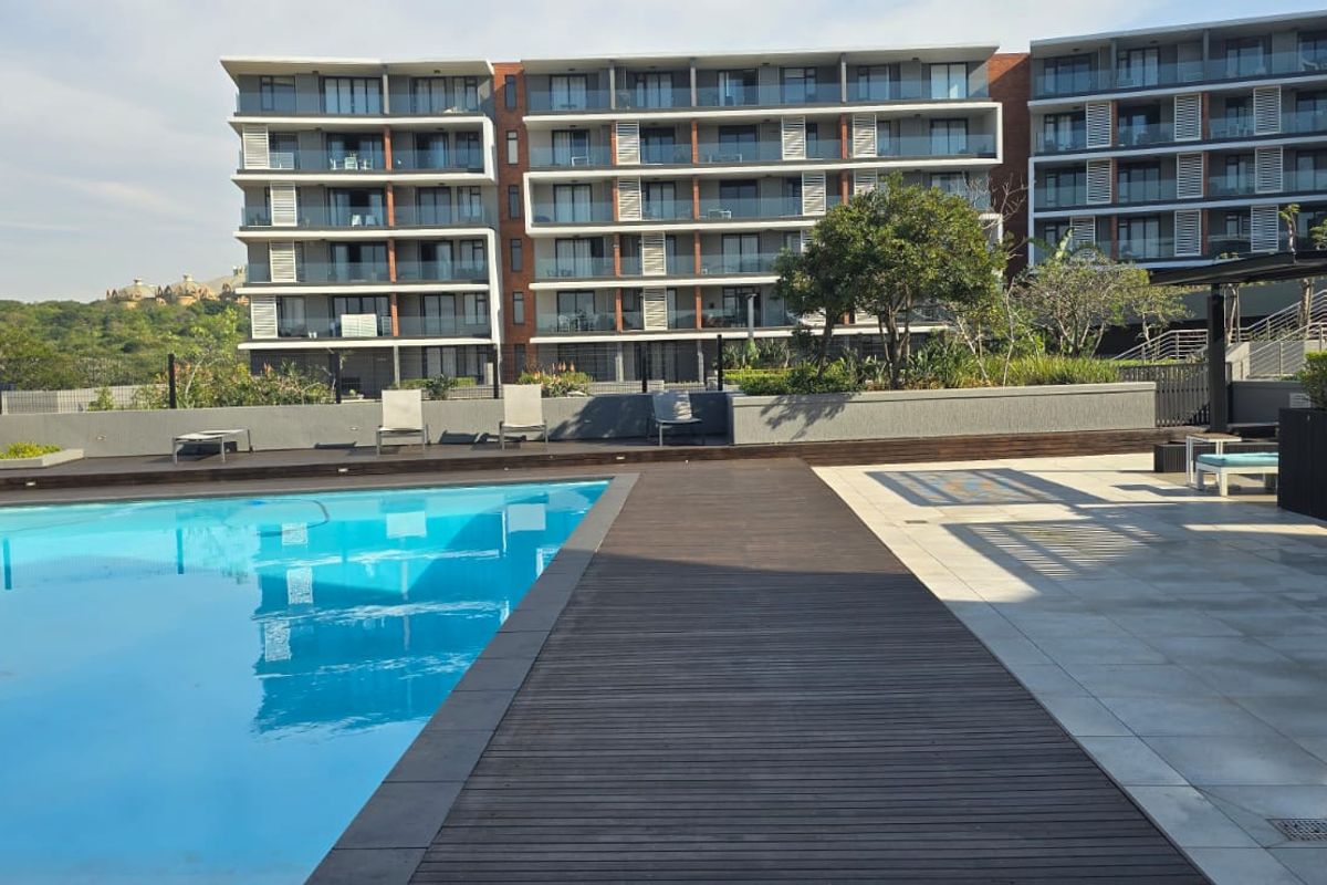 A well-maintained communal swimming pool area featuring timber decking, tiled patio space, and modern glass balustrades set against a contemporary apartment building.