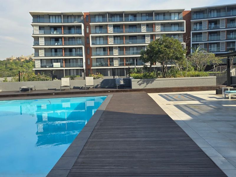 A well-maintained communal swimming pool area featuring timber decking, tiled patio space, and modern glass balustrades set against a contemporary apartment building.