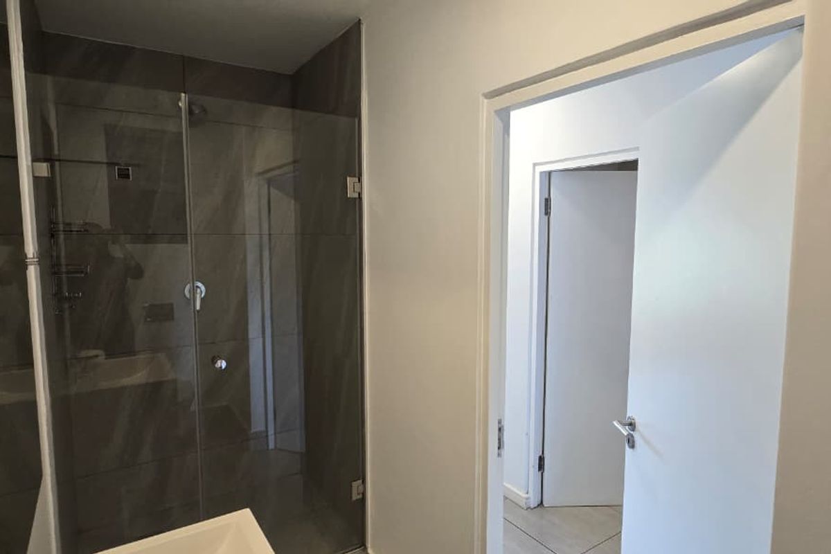 A modern bathroom featuring a glass-enclosed shower with dark tiling and neutral wall finishes, leading into a hallway.