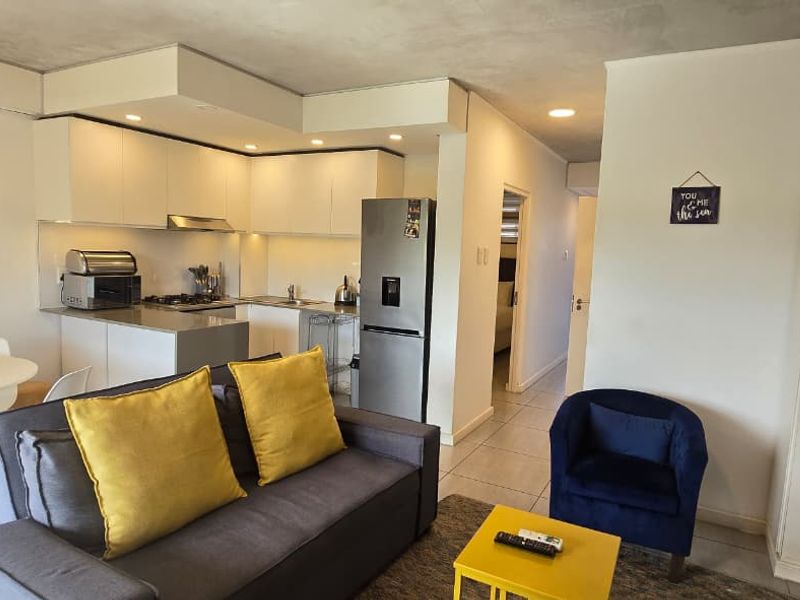 An open-plan living and kitchen area featuring modern white cabinetry, stone countertops, recessed lighting, and neutral tiled flooring throughout.