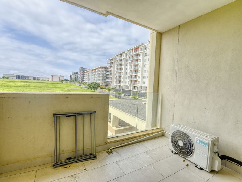 A covered balcony featuring tiled flooring, a glass balustrade, and an external air conditioning unit, offering an open view of the surrounding apartment complex.