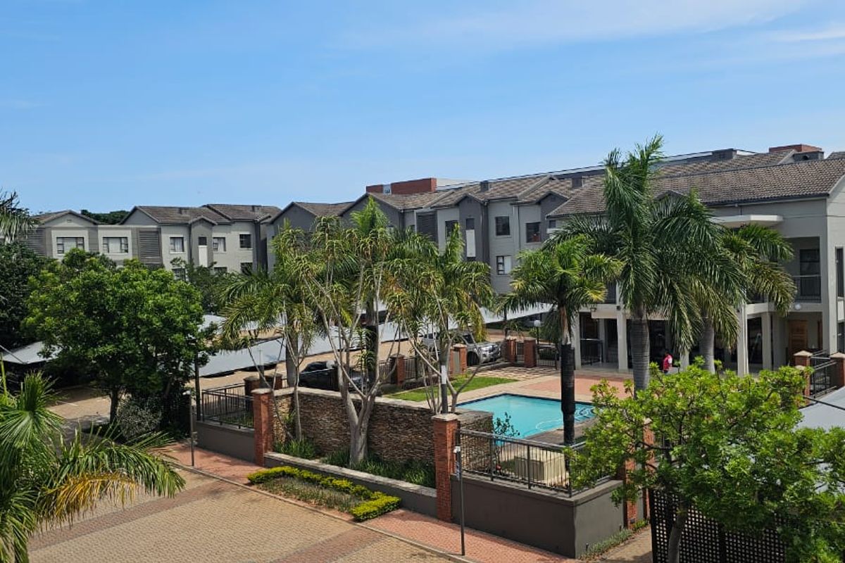 An elevated view of the apartment complex grounds, featuring a swimming pool, paved parking areas with shade structures, and established landscaping.