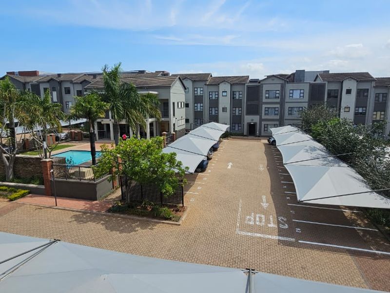 Aerial view of the apartment complex featuring a communal swimming pool, paved parking area with shade sail carports, and established landscaping.