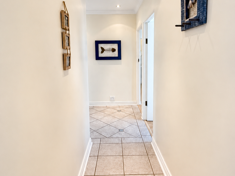 A clean and well-maintained hallway featuring neutral wall finishes, recessed downlighting, and durable ceramic floor tiles leading to adjacent rooms.