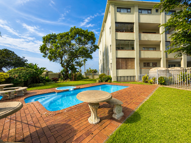 A well-maintained communal swimming pool area featuring brick paving, landscaped gardens, and perimeter fencing set against the apartment building facade.