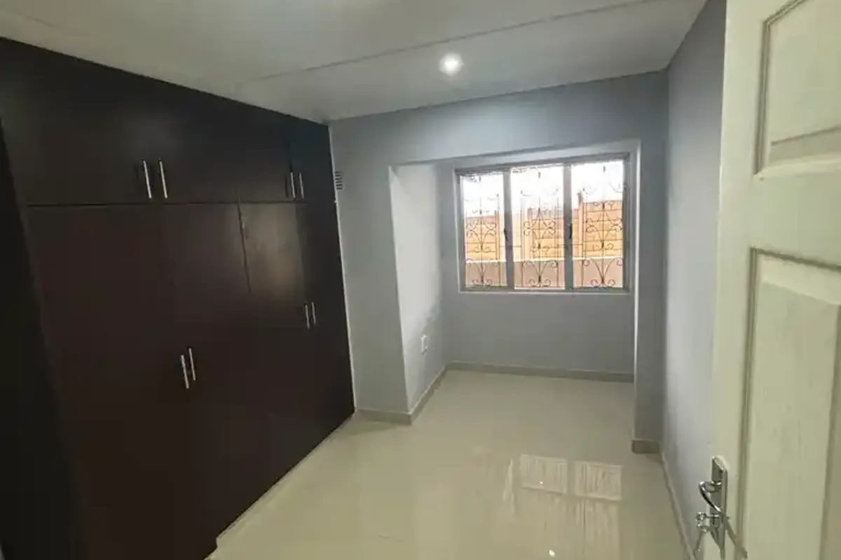 A clean, empty bedroom featuring dark wood built-in cupboards, polished tiled flooring, and a window fitted with security bars.