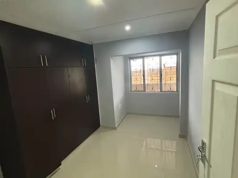 A clean, empty bedroom featuring dark wood built-in cupboards, polished tiled flooring, and a window fitted with security bars.