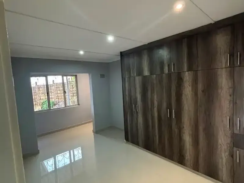 A bright, empty bedroom featuring modern wood-finish built-in cupboards, polished tiled flooring, and security bars on the windows.