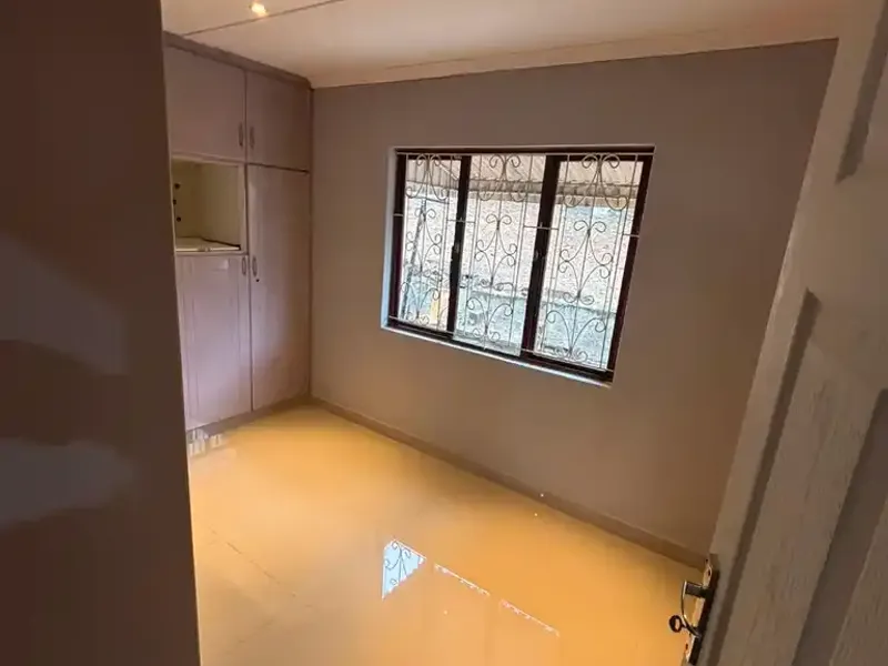 A clean, empty bedroom featuring neutral wall tones, polished tiled flooring, built-in storage cupboards, and secure window burglar bars.