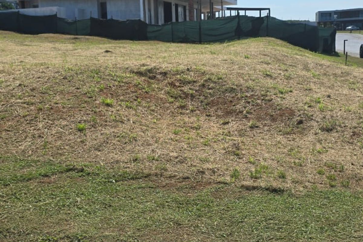 A view of a vacant residential plot featuring a sloped landscape and an unfinished modern building structure in the background.