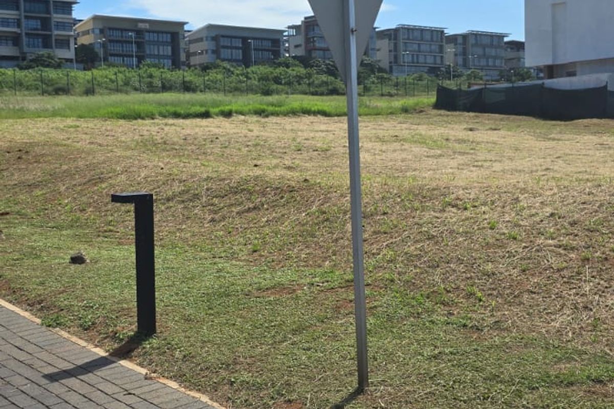 A view of a vacant residential plot featuring a paved walkway, a bollard light, and a traffic sign, with surrounding residential development in the background.