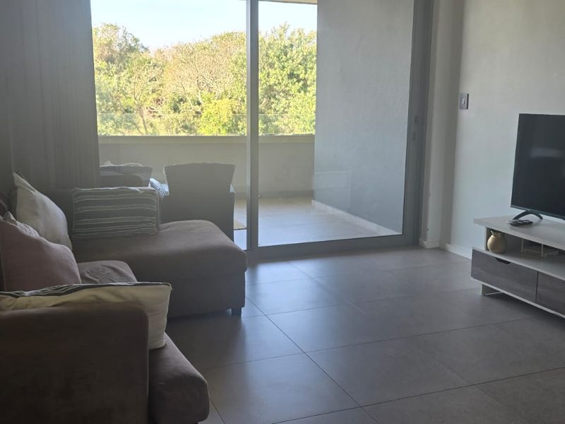 A bright living room featuring large sliding glass doors that open onto a balcony, with neutral tiled flooring throughout.