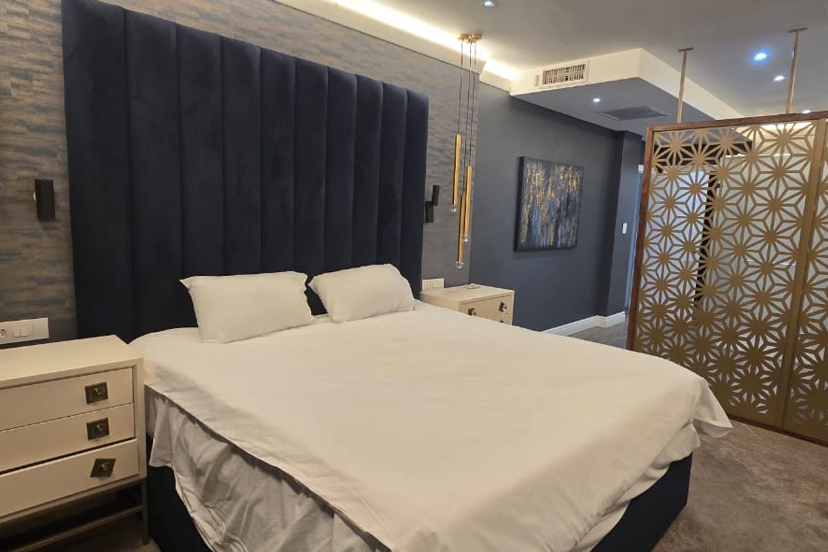 A modern, well-appointed bedroom featuring a large upholstered headboard, recessed ceiling lighting, ducted air conditioning, and a decorative metal privacy screen.