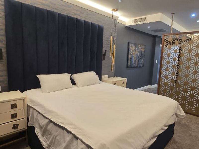 A modern, well-appointed bedroom featuring a large upholstered headboard, recessed ceiling lighting, ducted air conditioning, and a decorative metal privacy screen.