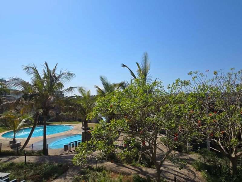 A view of the communal swimming pool area surrounded by lush tropical landscaping and mature trees within the apartment complex grounds.